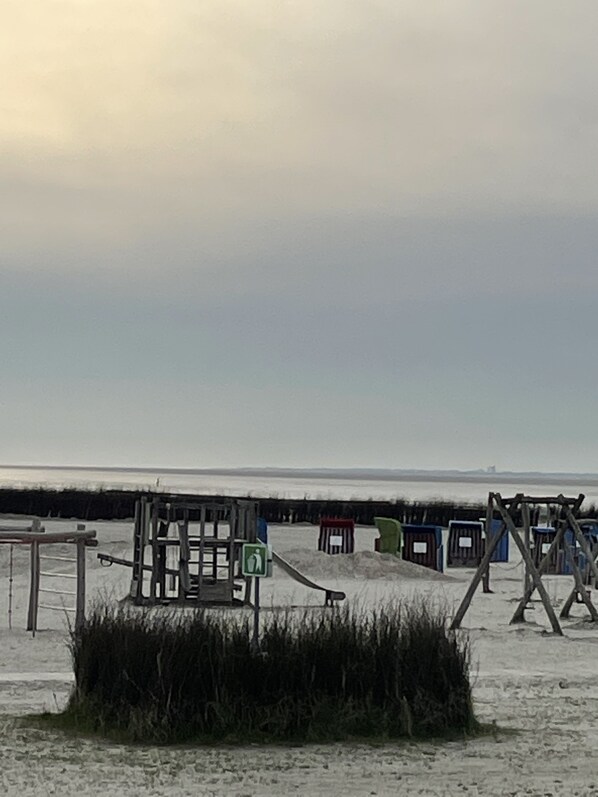 A sandy beach area with wooden structures, including a swing set and a small building with a green sign. - Küstenbadeort Neßmersiel Nordseeküste (26553 Neßmersiel)