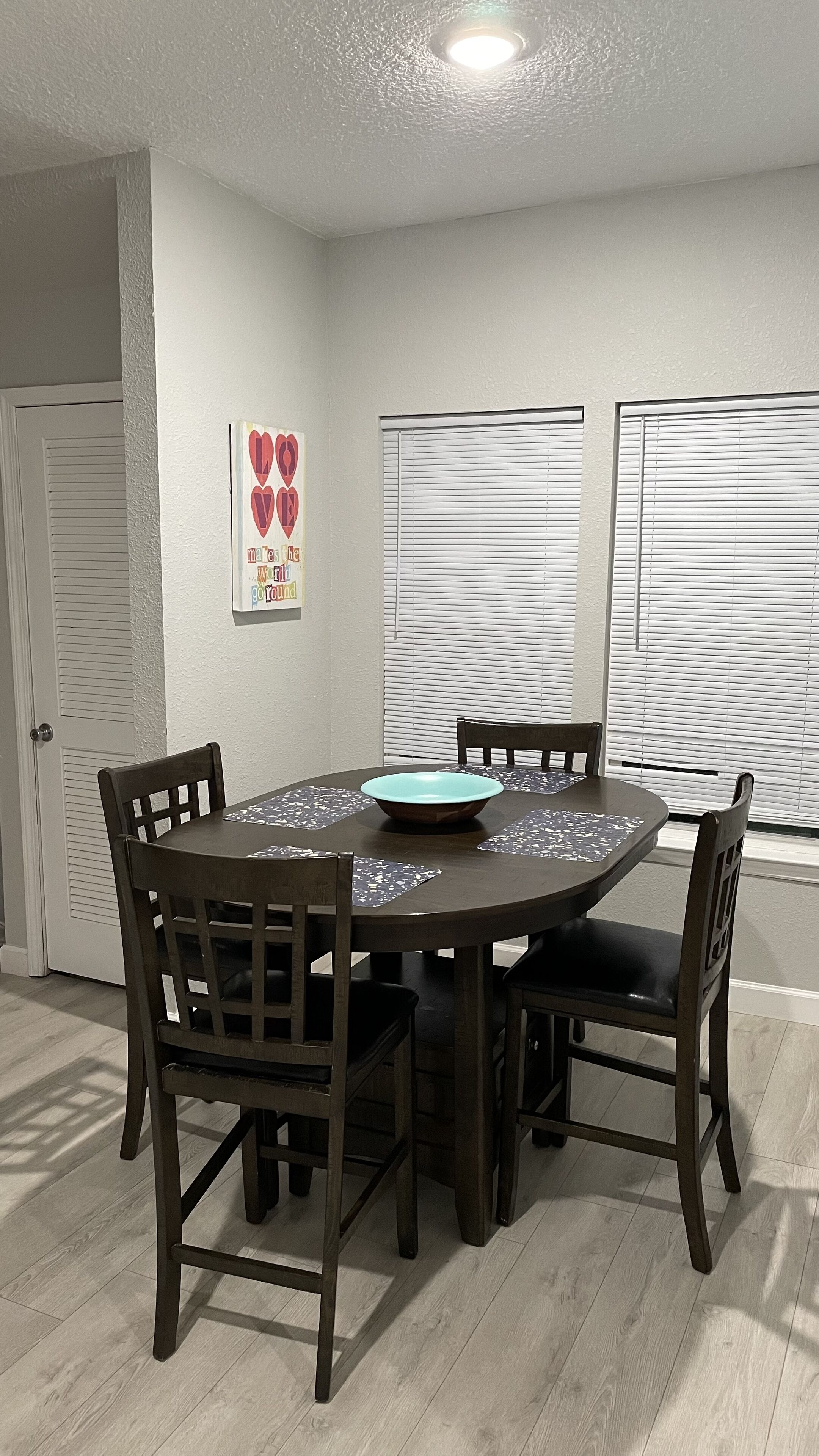 A dining area with a round table, four chairs, a teal bowl, and a wall decoration with hearts.