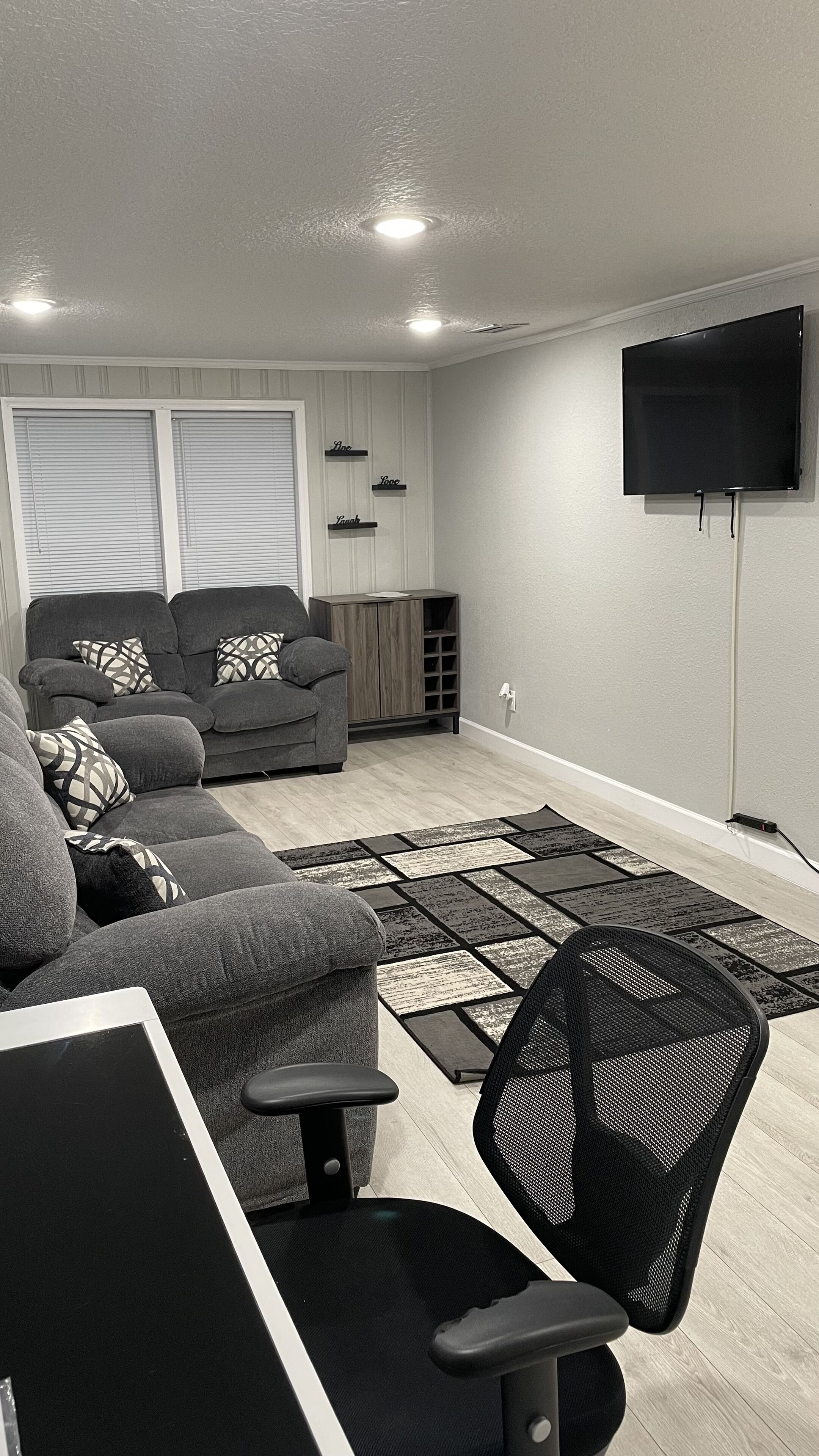 Ein modernes Wohnzimmer mit grauem Sofa, schwarzem Bürostuhl und einem flachbildfernseher an der Wand.