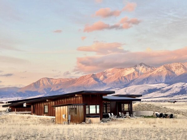 A modern wooden cabin with a flat roof, large windows, and a patio area with outdoor seating.