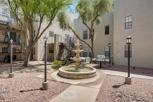 Stroll through the serene courtyard, surrounded by lush trees and a central water feature, with comfortable outdoor seating and a staircase to the upper level.