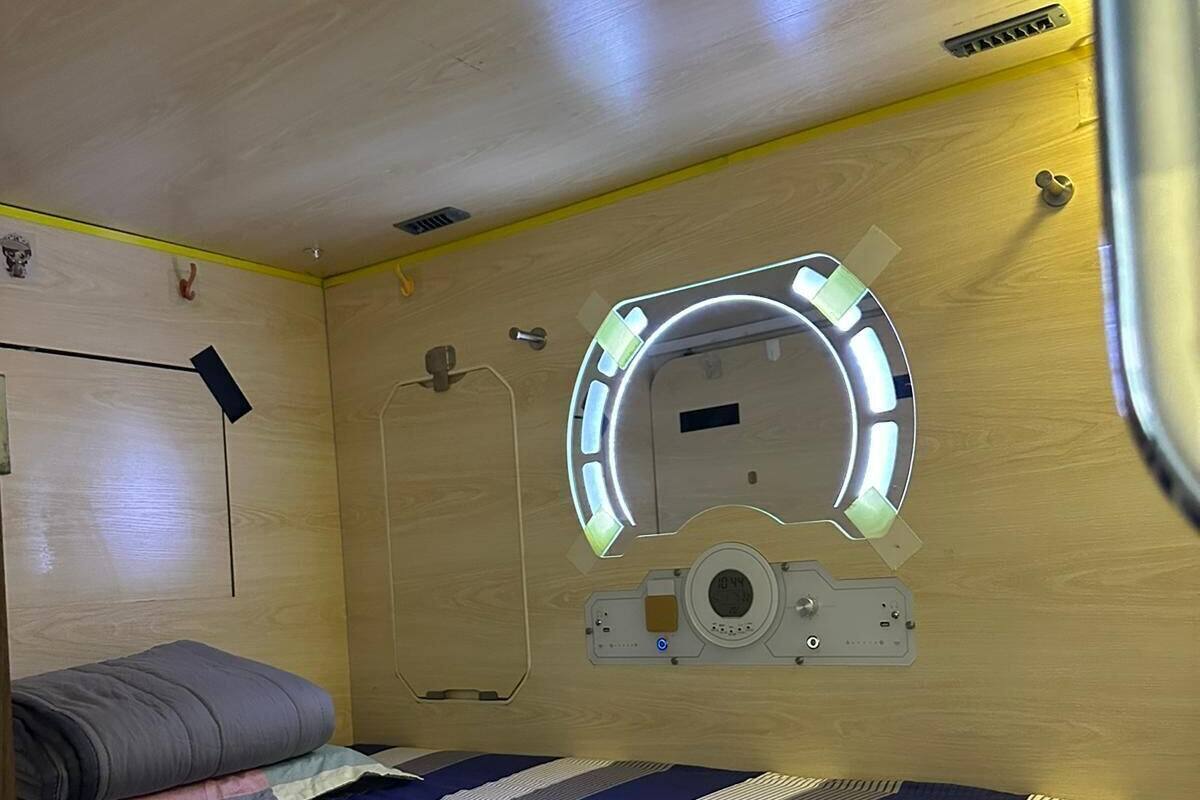 A compact room with a bed, a circular window with lights, a small control panel, and a door.
