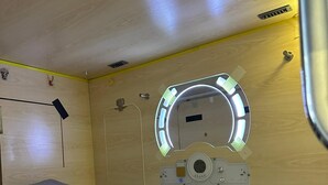 Une pièce de taille réduite comprenant un lit, une petite fenêtre avec un éclairage circulaire et un panneau de commande fixé au mur.