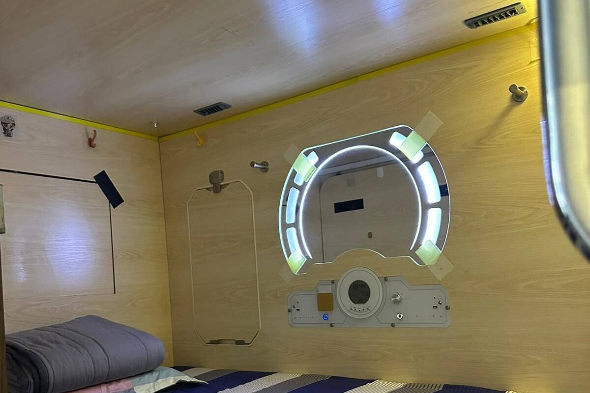A compact room with a bed, a small window with a circular light, and a wall-mounted control panel.