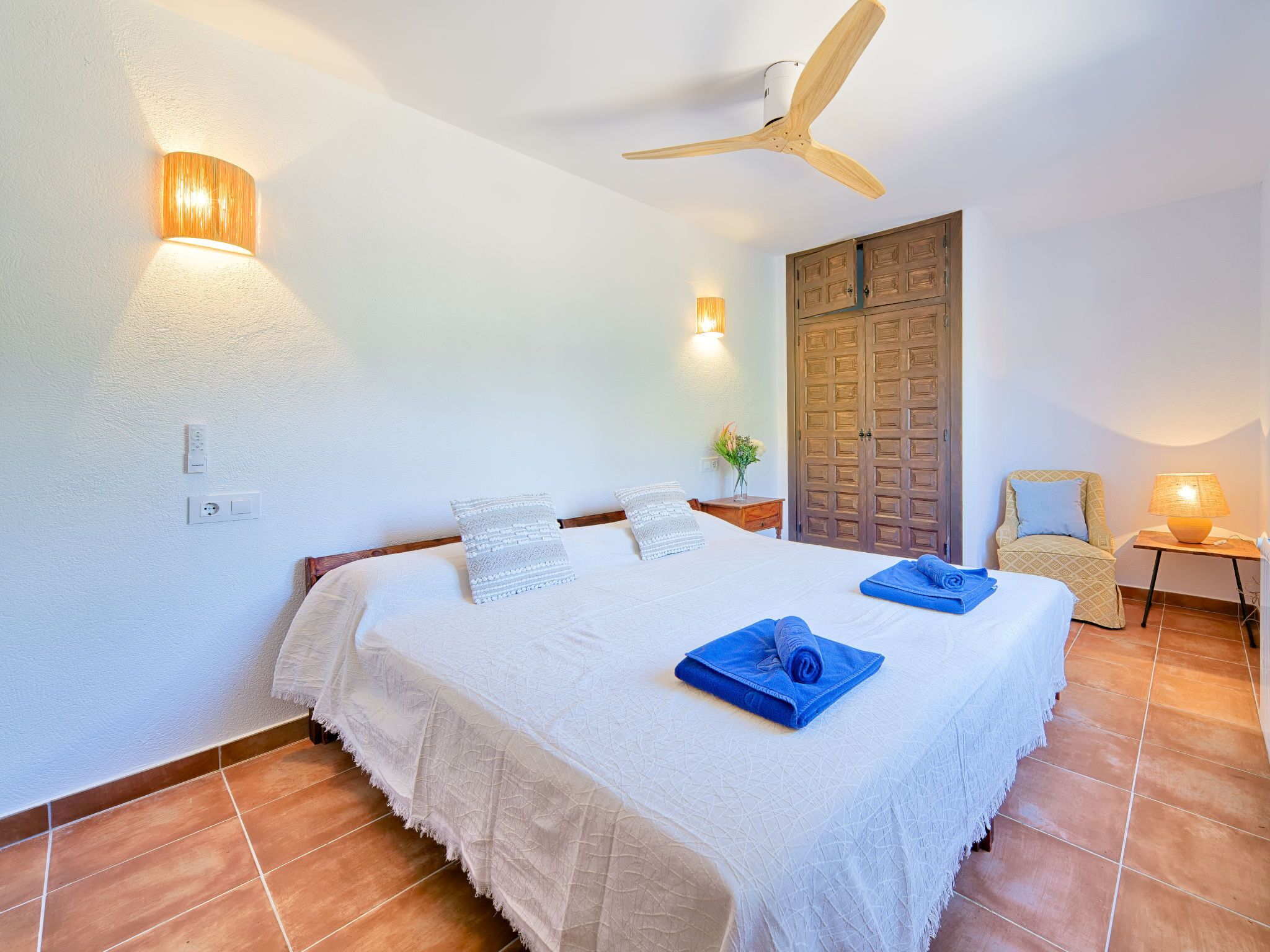 A bedroom with a bed, a ceiling fan, a wooden wardrobe, a chair, and a small table with a lamp.
