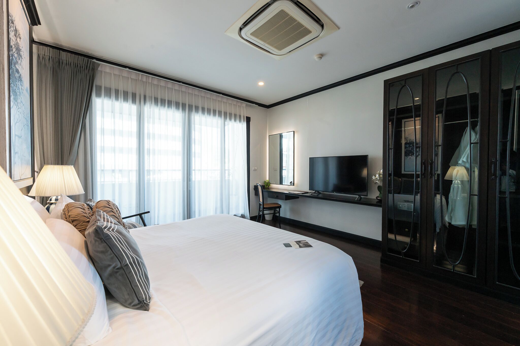 A hotel room with a large bed, a desk with a flat-screen TV, a chair, a wardrobe, and a window with curtains.