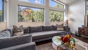 Un salon moderne avec un canapé gris, une table basse en verre agrémentée d’objets décoratifs et de grandes fenêtres offrant une vue sur les arbres et les montagnes.