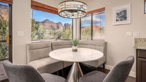 Un coin repas avec une table ronde, des sièges rembourrés, un luminaire suspendu, une plante en pot et une vue sur les montagnes à travers de grandes fenêtres.