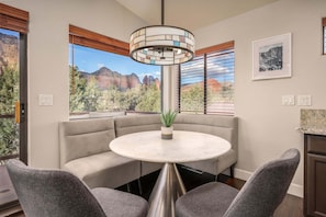 A dining area with a round table, cushioned seating, a hanging light fixture, a potted plant, and a mountain view through large windows.