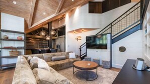 A spacious living area with a large sofa, a coffee table, and a TV. There is a staircase with a railing and a kitchen area in the background.