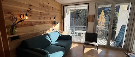 A modern living room with a wooden wall, a blue sofa, a small table, and a balcony with a view of trees.