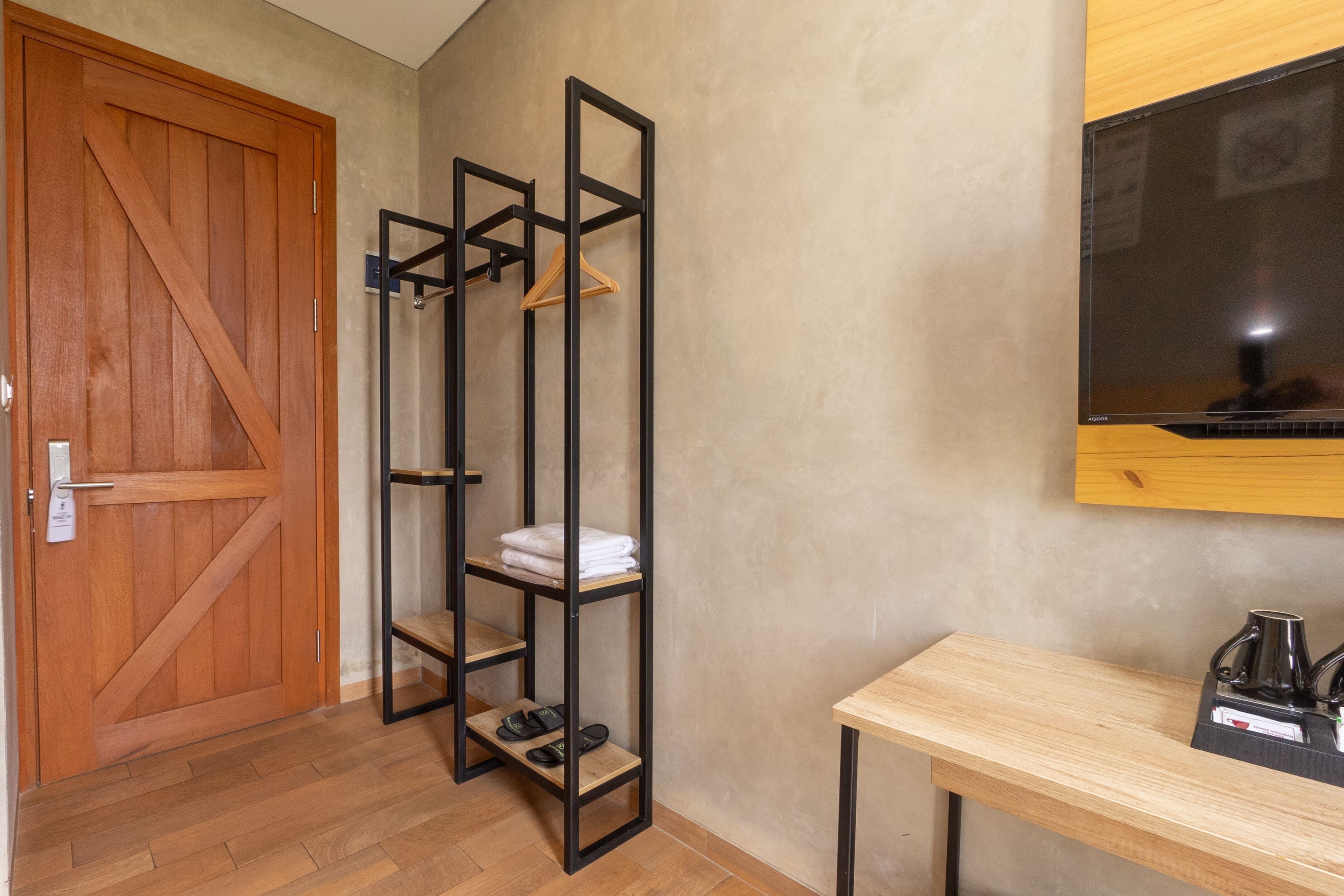 Une pièce avec une porte en bois, une étagère en métal noir où sont pliées des serviettes et rangées des chaussures, une table en bois avec une bouilloire, et un téléviseur fixé au mur.