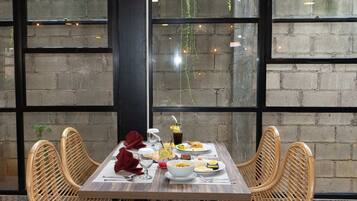 A dining table set with plates, glasses, and cutlery, surrounded by wicker chairs, with a brick wall and large windows in the background.