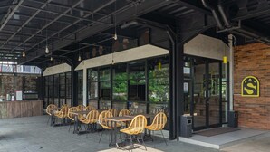 Un café al aire libre con sillas y mesas de mimbre, ventanas amplias y una pared de ladrillo con un logo.
