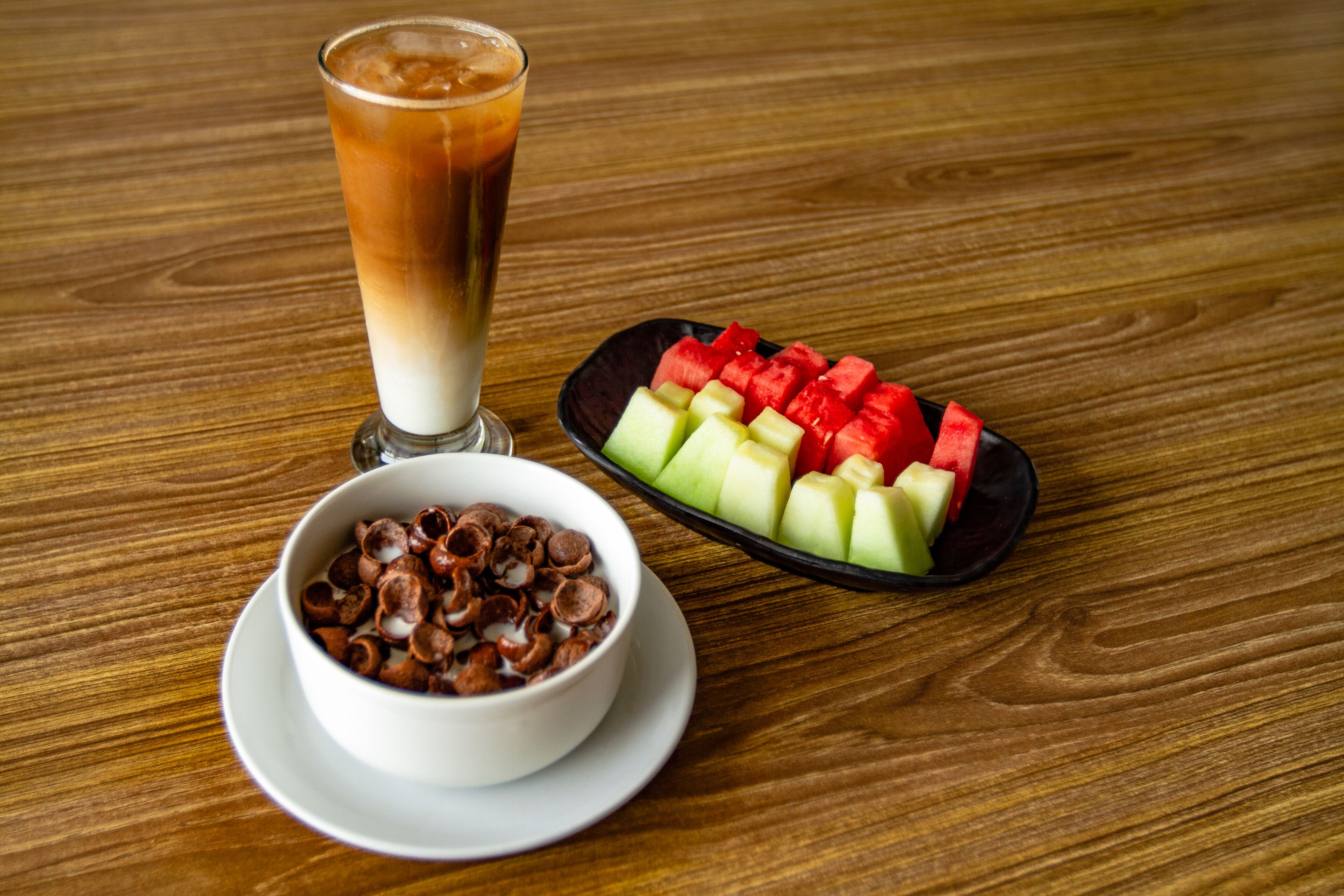 Una mesa de madera con un vaso de café helado, un tazón de cereales y un plato de fruta.