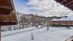 A wooden cabin with a snow-covered roof, a clear sky, and a view of a snowy landscape with trees and distant buildings.