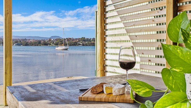Une table avec un verre de vin rouge, un morceau de fromage et des tranches de pain, le tout disposé devant une clôture en bois et avec en arrière-plan une vue sur un lac où navigue un voilier.