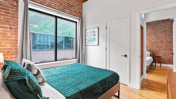 A bedroom with a brick wall, a large window, a bed with green bedding, and a door leading to another room.