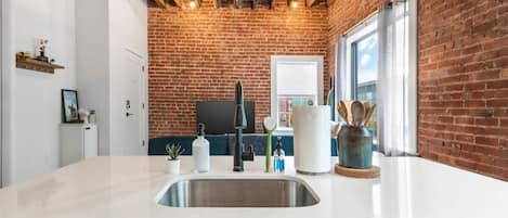 A kitchen with a white countertop, stainless steel sink, and a brick wall.