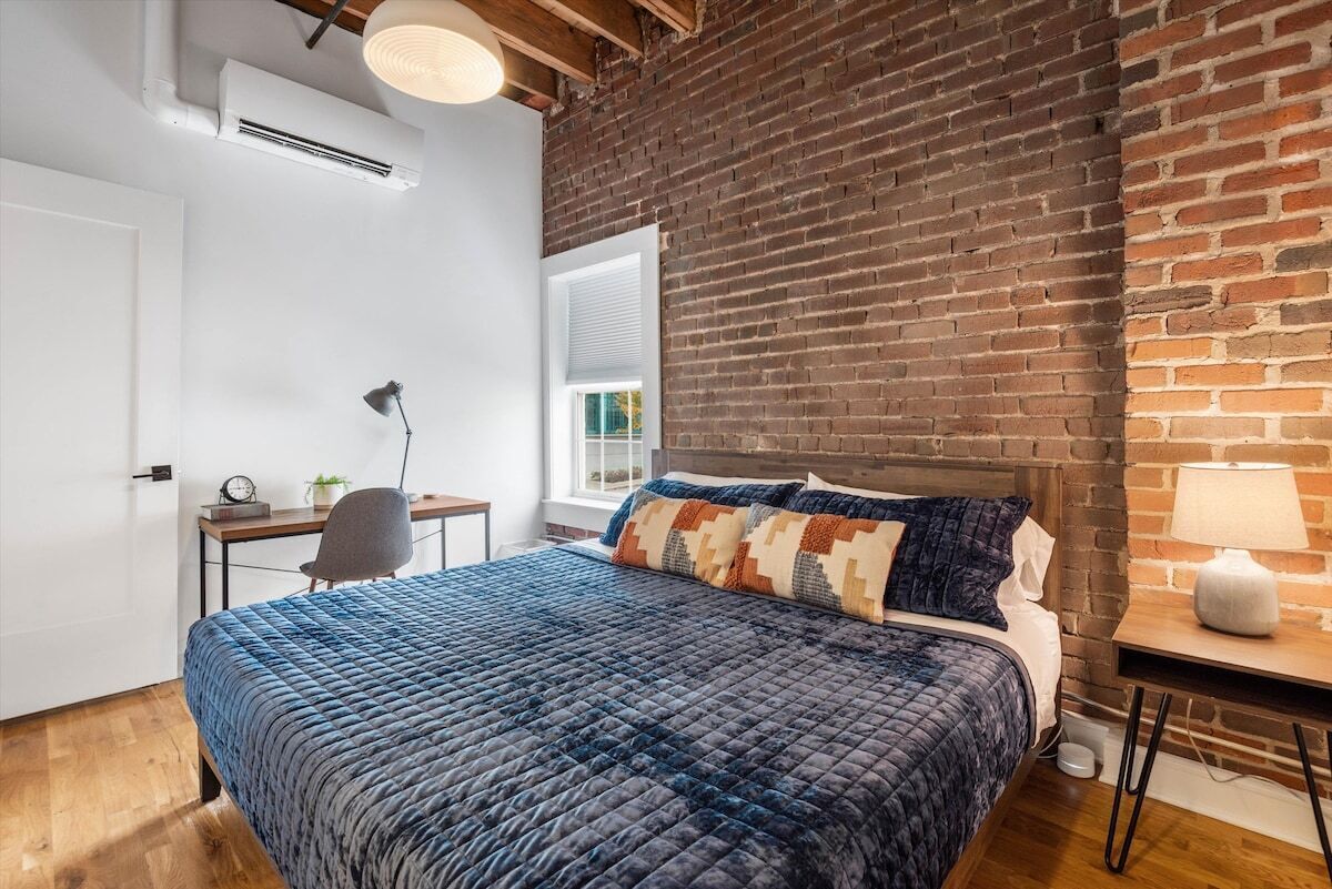 A bedroom with a brick wall, a bed with a blue quilt, a desk with a chair, and a window with blinds.