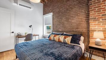 A bedroom with a brick wall, a bed with a blue quilt, a desk with a chair, and a window with blinds.