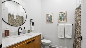 Une salle de bain avec un miroir rond, un lavabo blanc, des toilettes et un rideau de douche à motifs.