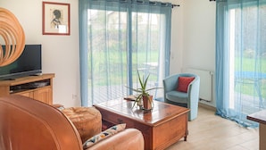 A living room with a sofa, coffee table, TV, and a chair with a red cushion. There are two windows with curtains, and a potted plant on the coffee table. - 3 bedroom stunning home in Trévou-Tréguignec (Trévou-Tréguignec)