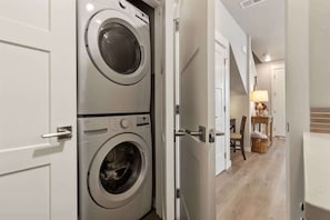 Uma lavanderia com duas máquinas de lavar empilhadas, uma porta que dá para um corredor e uma cesta de vime no chão.