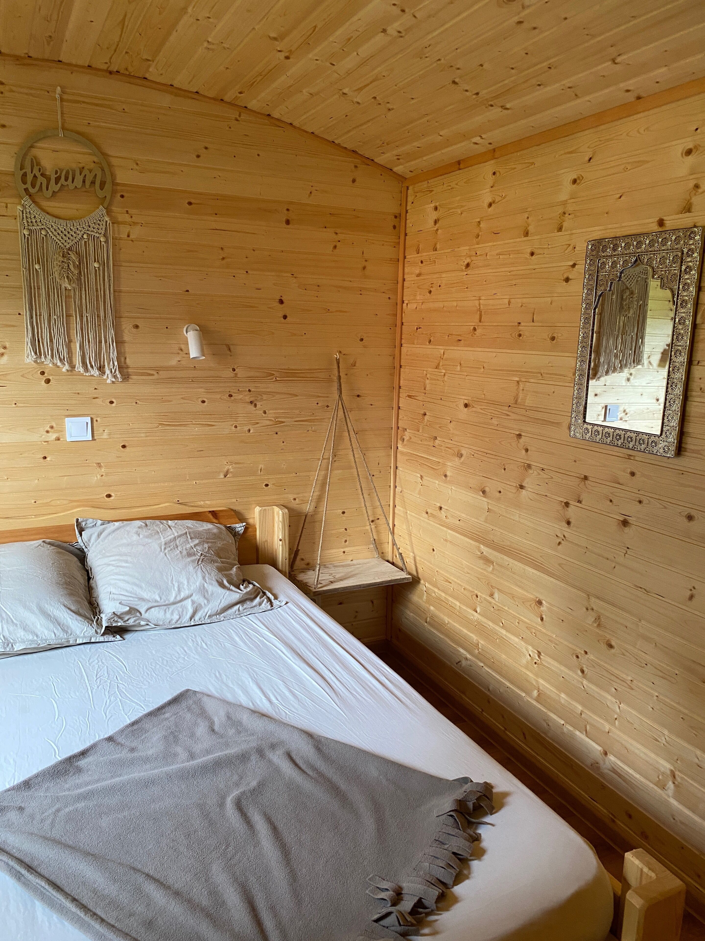 A wooden cabin room with a bed, pillows, a macrame wall hanging, a hanging chair, and a mirror.