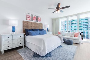 A bedroom with a large bed, white nightstands, a ceiling fan, and a view of a building through the window.