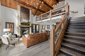 A spacious living room with a fireplace, a large brown sofa, a wooden staircase, and a high ceiling with exposed beams. - Rocky Mountain Gem!  Fireplace and Private Jacuzzi, Two Large patios, (Estes Park)