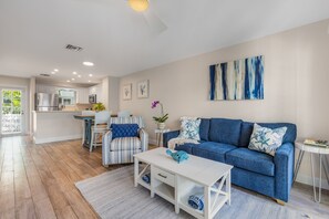 Un salon moderne avec un canapé bleu, une table basse blanche, un fauteuil à rayures et une cuisine en arrière-plan.