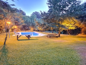 Uma área de piscina com mesa e bancos, cercada por árvores e iluminada por luzes.