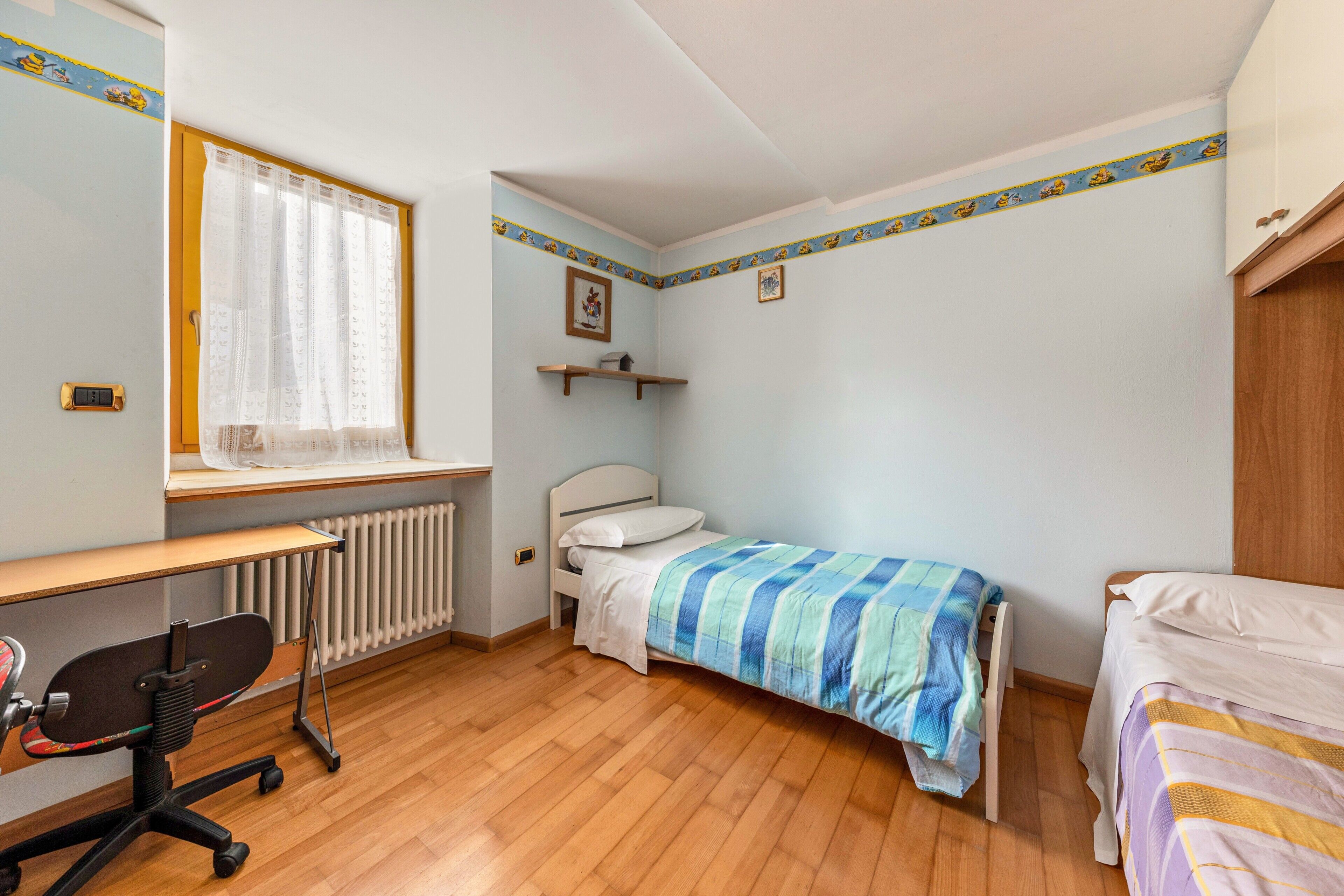 A bedroom with a wooden floor, a desk with a chair, two beds, a radiator, and a window with curtains.