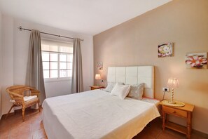 A bedroom with a bed, a chair, a nightstand, a lamp, and a window with curtains.