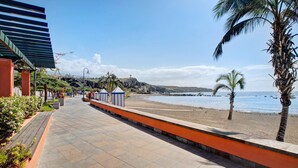 Ein gepflasterter Weg führt zu einem Strand mit Palmen, einer felsigen Klippe und blauem Himmel.