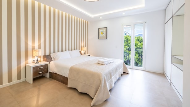 Une chambre moderne avec un grand lit, une table de chevet avec une lampe, un tableau encadré au mur et une vue sur la verdure par la fenêtre.
