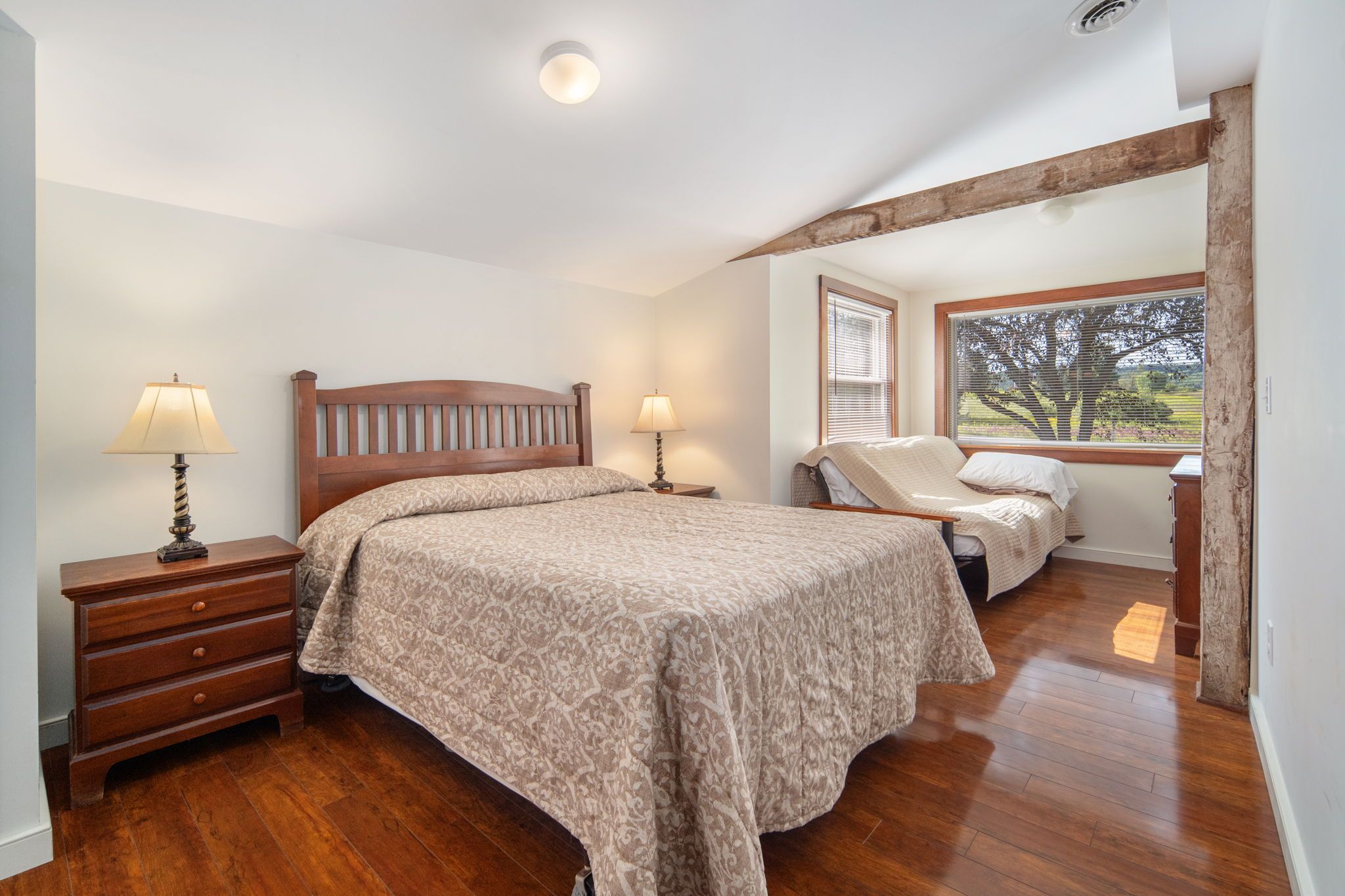 A bedroom with a wooden bed, two bedside tables with lamps, a dresser, and a window with blinds.