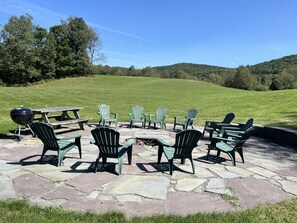 An outdoor seating area with Adirondack chairs and a grill, set on a stone patio with a view of a grassy hill and trees.