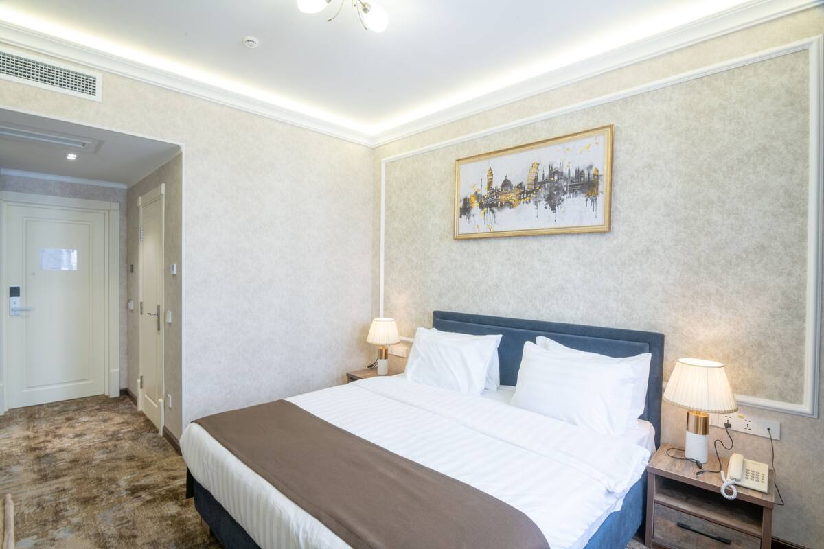 A hotel room with a large bed, two bedside tables with lamps, a wall-mounted artwork, and a door to another room.
