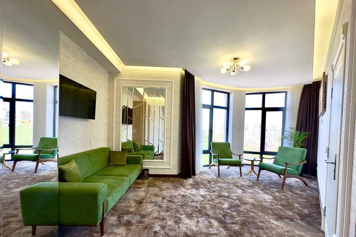 A spacious room with a green sofa, multiple chairs, a large mirror, and a view through large windows.