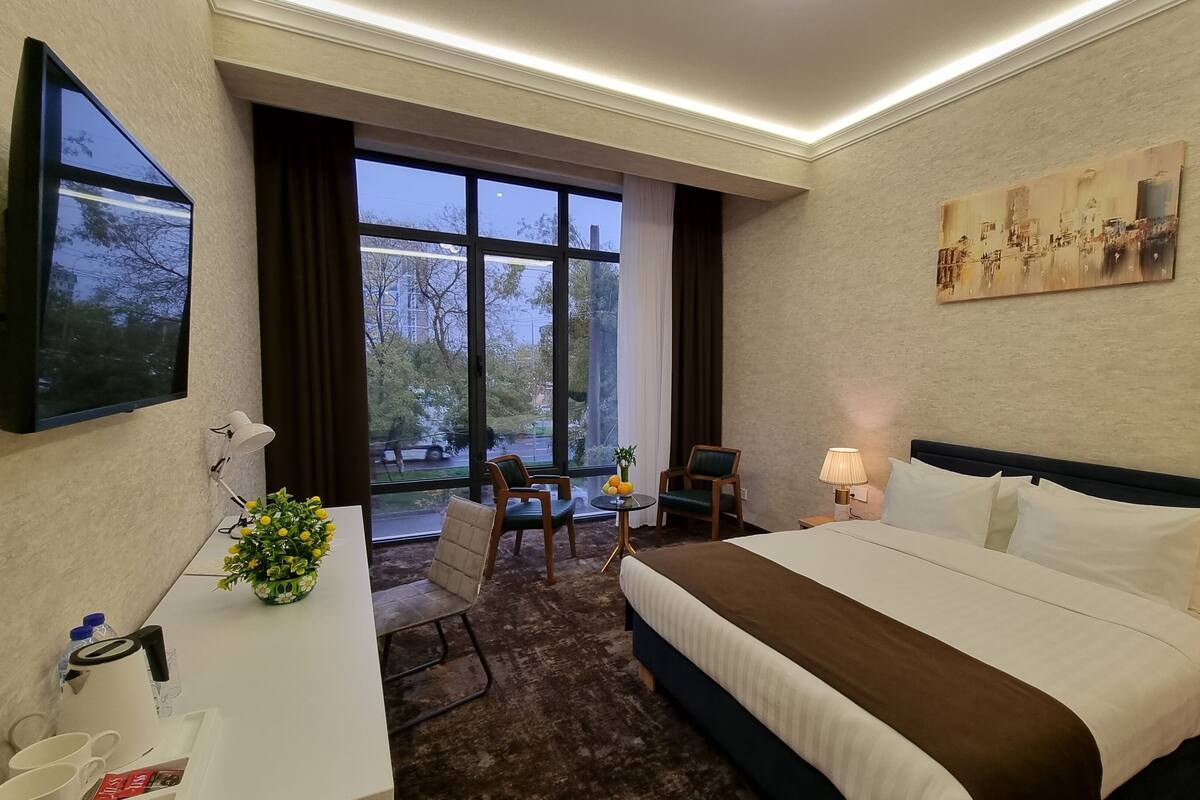 A hotel room with a large bed, a desk with a lamp, a chair, a small table with fruit, and a painting on the wall.