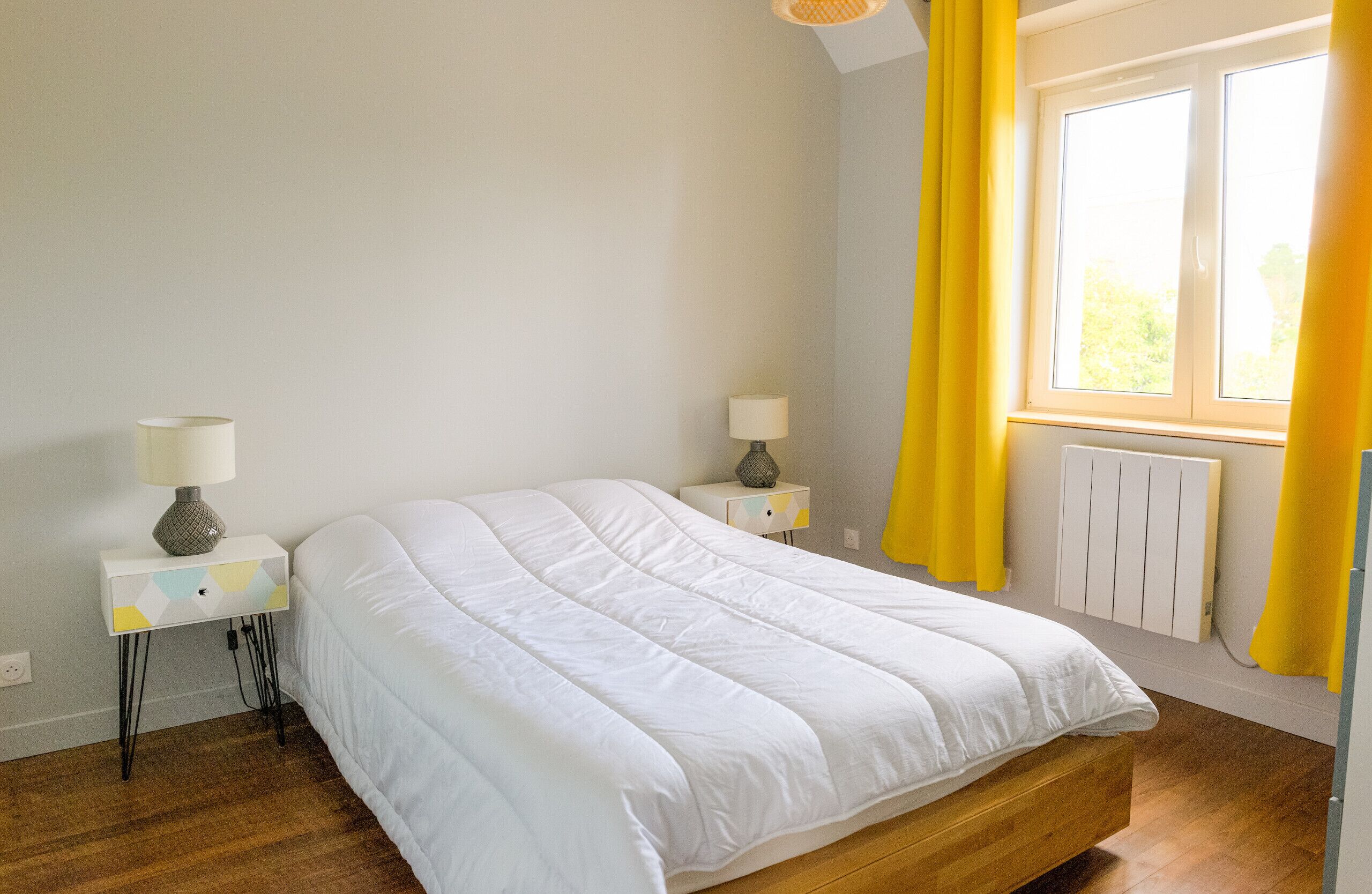 Une chambre à coucher avec un lit, deux tables de chevet avec des lampes, une fenêtre avec des rideaux jaunes et un radiateur.