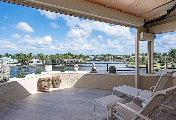 Un espace extérieur abrité, avec vue sur un lac, des palmiers et une statue. On y trouve des chaises longues et une table.