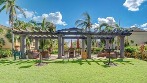A covered outdoor seating area with a pergola, chairs, and a small table, surrounded by grass and palm trees. - First Floor Corner Unit Condo On a Coveted Quad in Paseo of Fort Myers (Fort Myers)