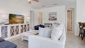 A spacious living room with a ceiling fan, a flat-screen TV, a white sofa, and a coffee table. - First Floor Corner Unit Condo On a Coveted Quad in Paseo of Fort Myers (Fort Myers)