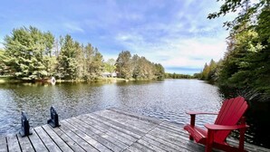 Une terrasse en bois avec un fauteuil Adirondack rouge et un barbecue noir, donnant sur un lac paisible entouré d’arbres.