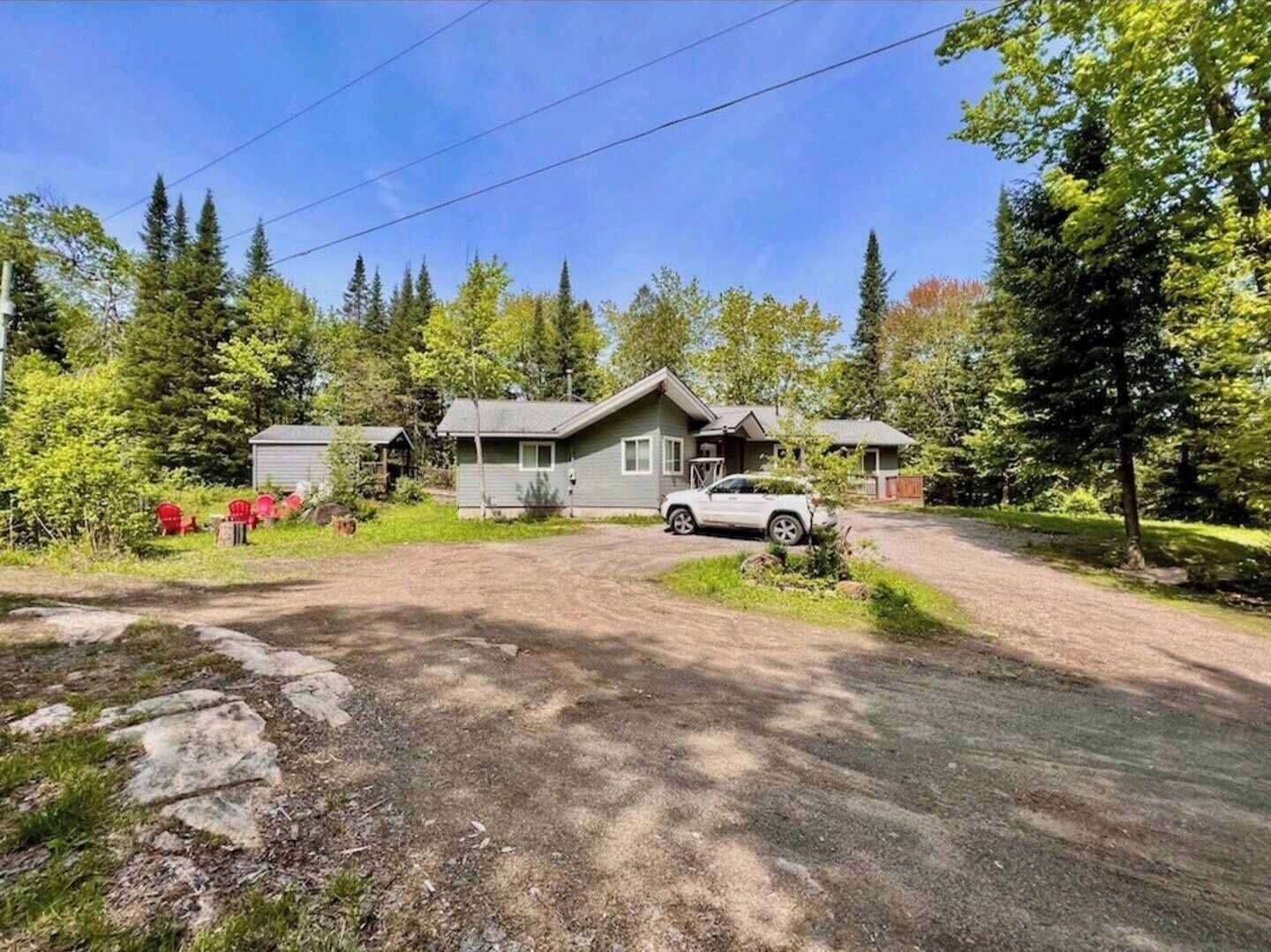 A two-story cabin with a gravel driveway, surrounded by trees and a small garden.