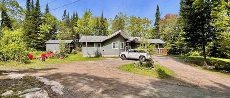 A two-story cabin with a gravel driveway, surrounded by trees and a small garden.