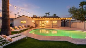 Un patio trasero con piscina, sillas de descanso y luces colgantes.